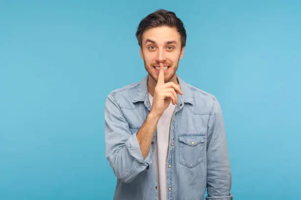 Please, keep quiet, this is confidential! Portrait of happy man in worker denim shirt showing silence gesture with finger on lips, asking for secrecy. indoor studio shot isolated on blue background