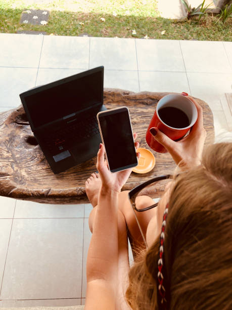 Modern freelance young woman using cellphone / laptop and drinking coffee on a terrace sofa.