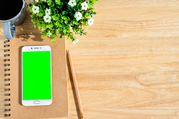 istock Smartphone green screen on notebook,flower pot tree,a pencil and coffee cup on wooden background,Top view with copy space office table. 1269879942