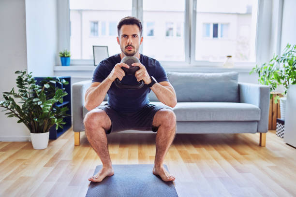 Home workout, young man doing squats with kettlebell in his apartment Home workout, young man doing squats with kettlebell in his apartment squatting position photos stock pictures, royalty-free photos & images