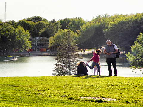 Family enjoying a peaceful evening by Montreal's Lac des castors park on the Mount Royal. A young girl is skipping rope while a man looks at his phone and the others are sitting on the lawn, looking at the view.