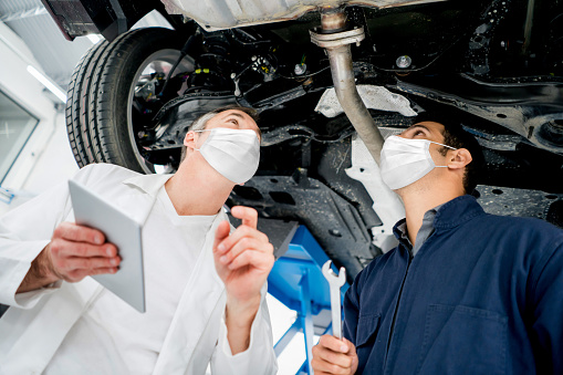 Mechanics working at an auto repair shop wearing facemasks and looking at a car's chassis â illness prevention concepts