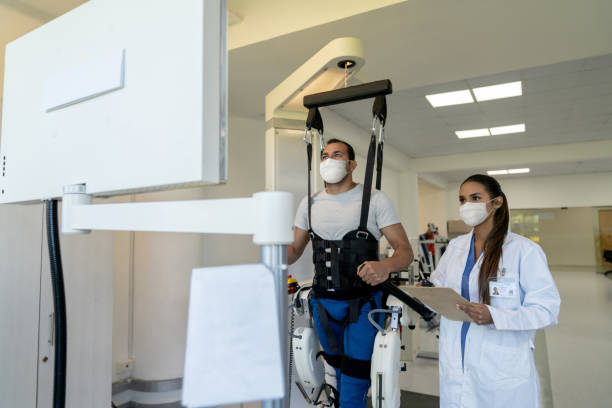 Patient doing physiotherapy with an exoskeleton with his physical therapist supervising and both wearing facemasks Patient doing physiotherapy with an exoskeleton with his physical therapist supervising and both wearing facemasks â pandemic lifestyle concepts powered exoskeleton photos stock pictures, royalty-free photos & images