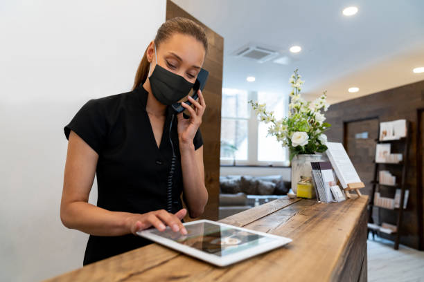 電話で話している間、フェイスマスクを着用してスパで働く女性 - hotel reception hotel receptionist health spa ストックフォトと画像