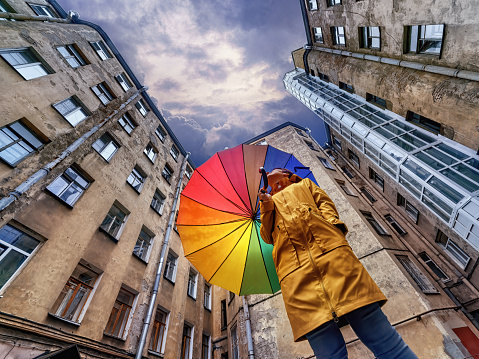 A woman in a bright yellow raincoat with a rainbow umbrella standing in an old courtyard well and looking at the dark stormy sky. Saint Petersburg, Russia