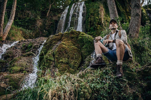 One man, male hiking alone in nature, he is sitting by the waterfall, holding bottle of water.