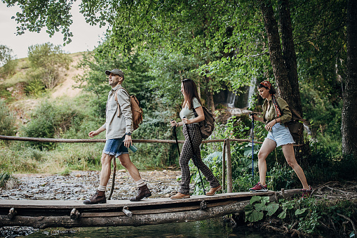 One man and two women, three friends hiking together in nature, they are crossing wooden bridge.