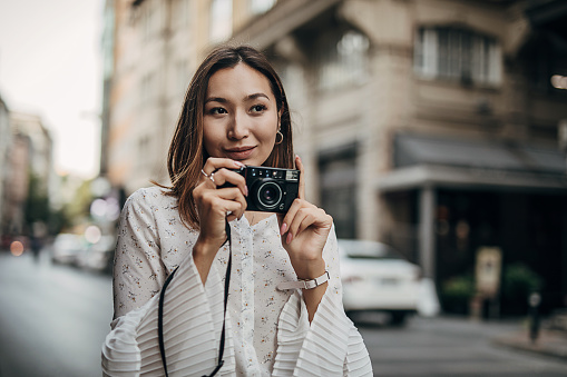 One woman, beautiful Asian woman standing downtown on the street, taking a pictures with camera.