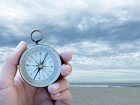 A woman holds a compass in her hand with a cold and dreary seashore in the background.