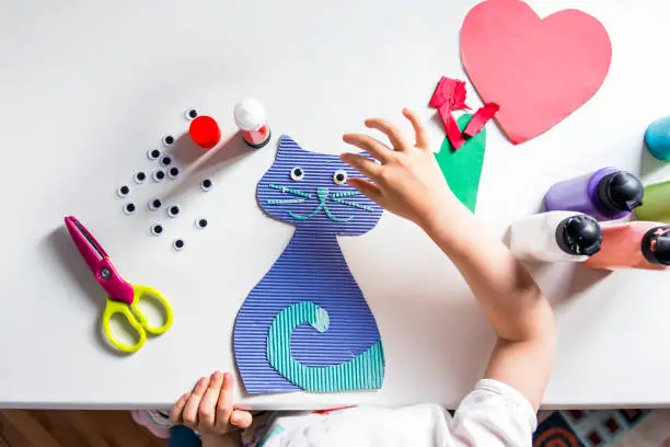 An unrecognizable little girl sticks googly eyes on a handcrafted cat figure with a glue stick.  Preschool paper and glue crafts activities. DIY