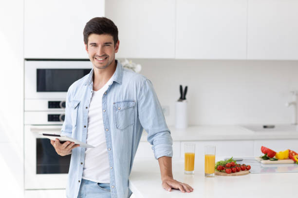 istock Young man standing on domestic kitchen holds digital tablet searching recipes for cook vegetable salad healthy food. 1269949169