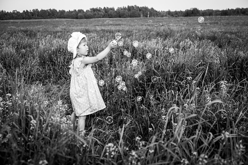 Young girl in dress walking at field and and blowing bubbles in black white colors. Summer time.