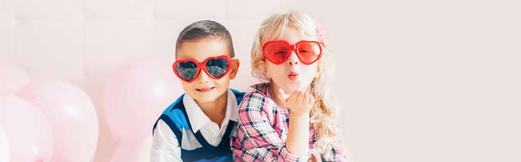 Valentine day holiday celebration. Web header banner for website. Two happy Caucasian cute funny children wearing heart shape sunglasses. Boy and girl hugging. Love, friendship and fun.