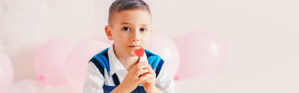 feliz menino caucasiano pensativo comendo doces de pirulito vermelho em forma de coração. o conceito de feriado do dia dos namorados. cabeçalho de banner da web para site. - child valentines day candy eating - fotografias e filmes do acervo