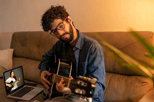 Latin man with beard practicing guitar lessons through video tutorial.