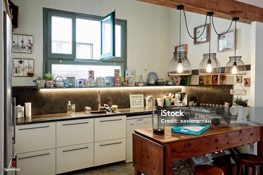 Eclectic Style Domestic Kitchen in Spanish Home Wide angle view of domestic kitchen with open window, natural light, and wooden table with stools for informal dining. Eclectic Style Stock Photo