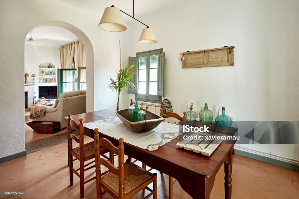 Dining Room with View into Living Room in Spanish Home Wide angle view of vintage dining table for four, hanging light fixture, knick knacks, and terracotta tile flooring with view of living room through archway. Living Room Stock Photo