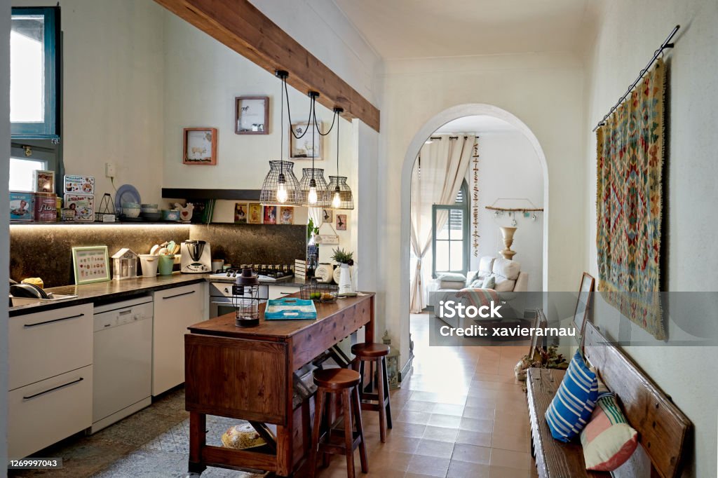 Rustic Domestic Kitchen in Spanish Home Wide angle view of kitchen with wooden counter-height dining table and stools, hanging light fixtures, bench, and view into living room. Rustic Stock Photo