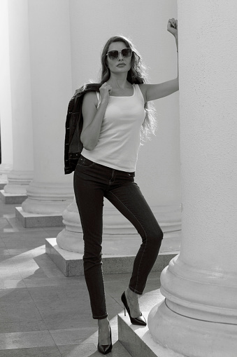 Black and white photo of a tall model in a white t-shirt and jeans. Stylish and beautiful girl, standing near the white columns.