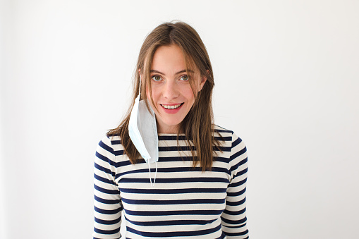 Delighted young female in casual striped shirt with protective mask hanging on ear looking at camera against white background