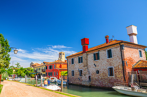 Torcello island with embankment promenade along water canal with motor boats and colorful buildings. Tower and blue clear sky background. Venetian Lagoon, Veneto Region, Northern Italy.