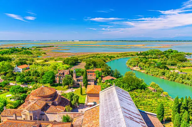 Aerial view of Torcello islands with buildings, water canal, swamp, green trees and bushes. Panoramic view of Venetian Lagoon from bell tower. Veneto Region, Northern Italy. Blue sky background.