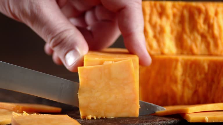 Male hand with a knife cut cheddar cheese into pieces.