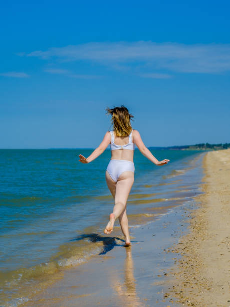 vista posteriore di una ragazza che scappa in lontananza lungo una spiaggia sabbiosa di mare spruzzi di acqua di mare. la ragazza indossa un costume da bagno bianco e occhiali da sole. libertà. fitness. giornata di sole estivo. spazio di copia - pin up girl foto e immagini stock