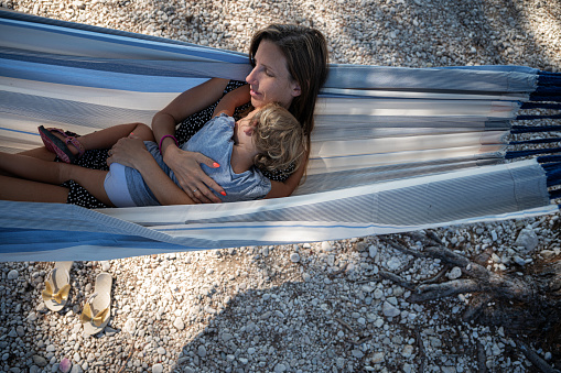 Top view of young mother and her toddler daughter cuddling and resting in a hammock in the shade on a sunny summer day.