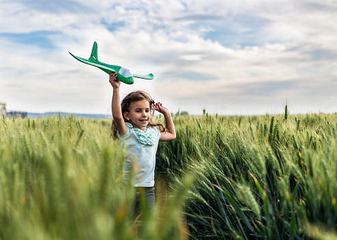 Happy little girl is pretending to be an airplane, while running through green wheat field. She is holding airplane toy up in the air.