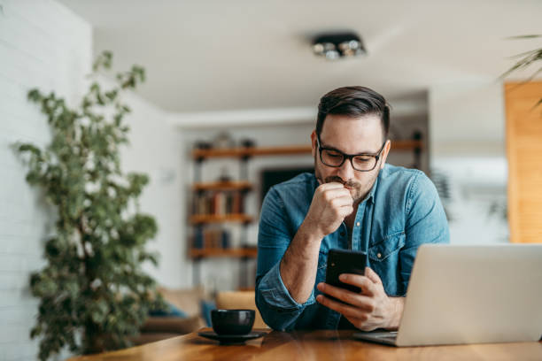 Portrait of a puzzled man looking at smart phone, sitting at table at home. Portrait of a puzzled man looking at smart phone, sitting at table at home. waiting stock pictures, royalty-free photos & images