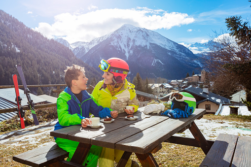 Mother look at and talk to a little boy in ski outfit sit, enjoy lunch break over mountain view after skiing