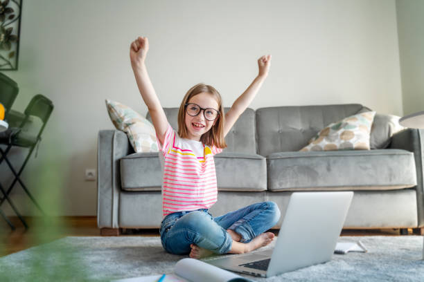 happy cute little girl 8 years old in a striped t-shirt and jeans with glasses sits at home on a carpet in front of a laptop, remote education technologies homework - 12 13 years fotos imagens e fotografias de stock