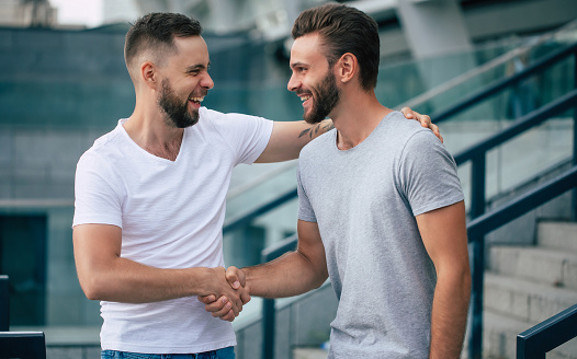 Two happy excited handsome bearded best friends in casual clothes are handshaking, hugging while standing outdoor.