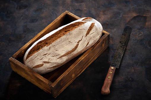 Wholegrain rye bread loaf in wooden box tray over dark wooden background with vintage knife