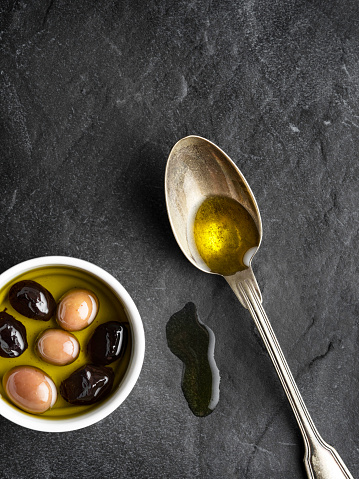 Cooking oil in bowl, olives and leaves on black textured table, flat lay. Space for text