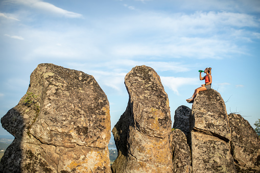 Action photo of extreme sport athlete woman trail runner sitting on huge rock and drinking water while taking a break on her running and climbing mountain cliff training. Extreme terrain.