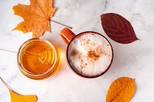 Top view of coffee with milk foam and cinnamon, jar of honey and orange autumn leaves on white marmor background
