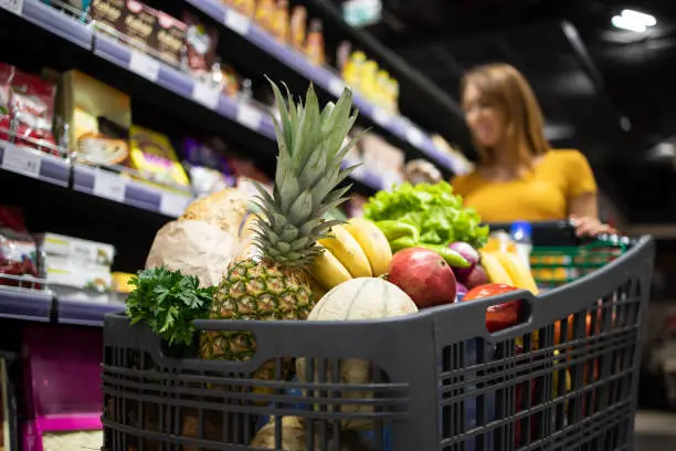 Supermarket shopping. Close up view of shopping cart overloaded with food while in background female person choosing products.