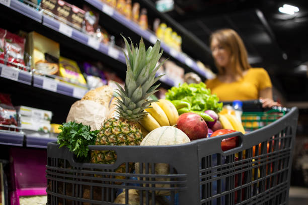 zakupy w supermarkecie. zbliżenie na koszyk przeładowany jedzeniem, podczas gdy w tle kobieta wybiera produkty. - pełny zdjęcia i obrazy z banku zdjęć