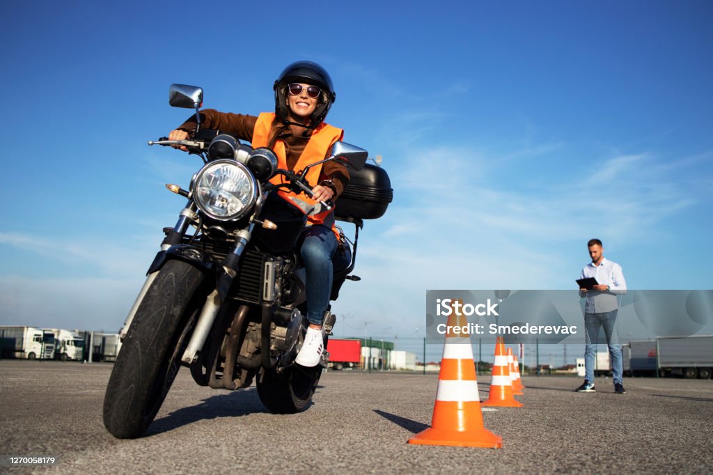 Female student with helmet taking motorcycle lessons and practicing ride. In background traffic cones and instructor with checklist rating and evaluating the ride. Motorcycle school of driving. Motorcycle Stock Photo