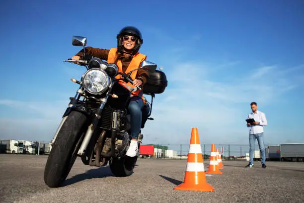 Photo of Female student with helmet taking motorcycle lessons and practicing ride. In background traffic cones and instructor with checklist rating and evaluating the ride. Motorcycle school of driving.