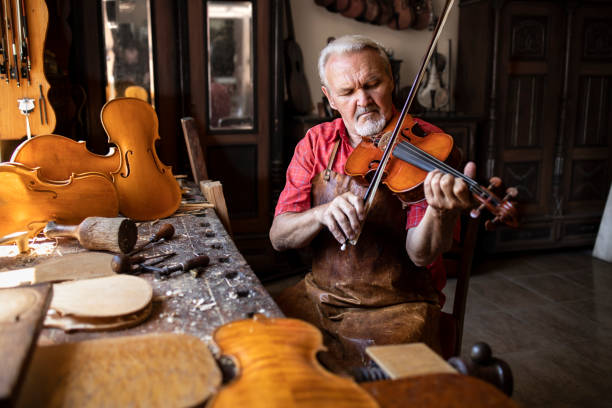 istock An elderly carpenter master checking quality of product. An experienced grey haired senior man woodworker wearing leather apron and sitting in his woodworking workshop playing violin. 1270059812