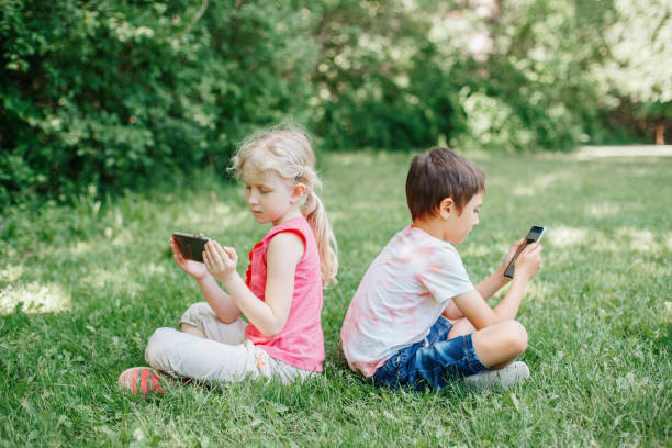 Boy and girl play games on smartphones outdoors. Kids digital gadget screen addiction. Children friends playing online sitting on grass in park. Contemporary problem of loneliness together. stock photo