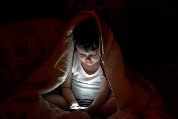 любопытный мальчик сидит в постели под белым одеялом и играет в игры на смартфоне. - ipad 3 ipad white caucasian стоковые фото и изображения