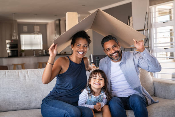Happy multiethnic family with child holding cardboard roof Portrait of smiling family sitting on couch holding cardboard roof and looking at camera. African and indian parents with funny daughter holding cardboard roof over heads while sitting on sofa in their new home. Happy mother and father with daughter in new house at moving day with copy space. covering stock pictures, royalty-free photos & images