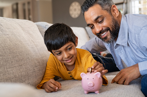 Happy indian son saving money in piggy bank with father. Lovely ethnic father teaching to little boy importance of saving money for future. Smiling middle eastern kid adding coin in piggybank while lying on couch with dad at home.