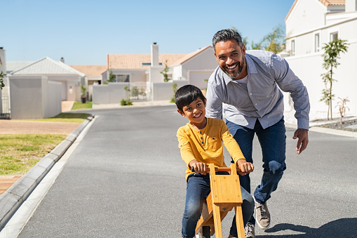 Happy latin father helping smiling boy to ride wooden balance cycle on street. Happy middle eastern child and young dad riding bike in lane. Smiling daddy teaching son to ride a balance bicycle, copy space.