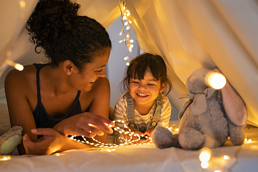 Playful mother and her daughter playing in children room in lighted tent. African mom and lovely cute girl having fun in kid hut at night at home. Mother and little daughter spending time together with strip lights.