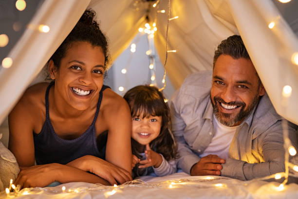 Happy parents with daughter have fun in cozy tent Portrait of little daughter lying with mother and father in illuminated tent. Cheerful man and happy woman lying in cozy hut with their cute daughter. Indian family lying on bed and having fun together. sheltering photos stock pictures, royalty-free photos & images